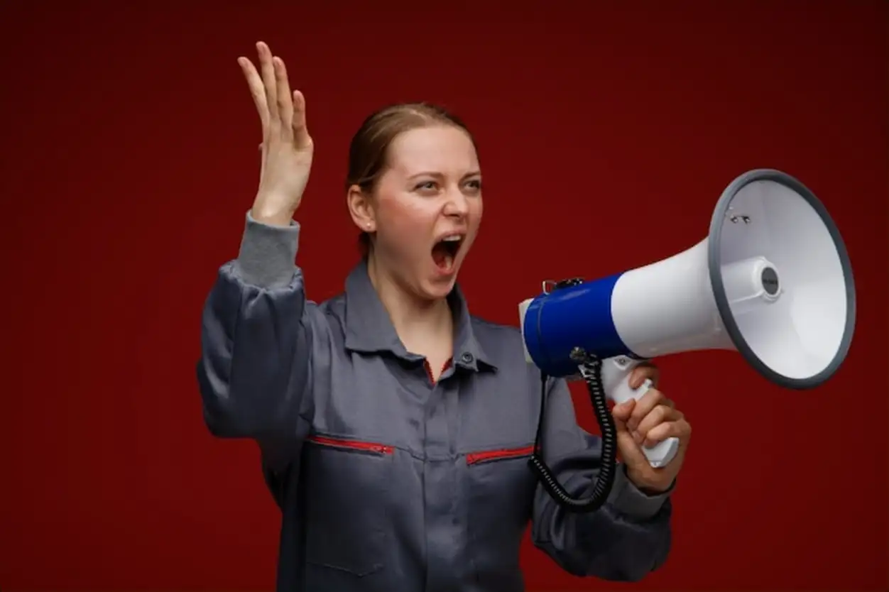Furious young blonde female engineer wearing uniform looking at side raising hand shouting in loud speaker